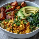 Colorful tofu scramble breakfast bowl with golden roasted sweet potatoes, fresh spinach, fluffy quinoa, and sliced avocado for a healthy start.